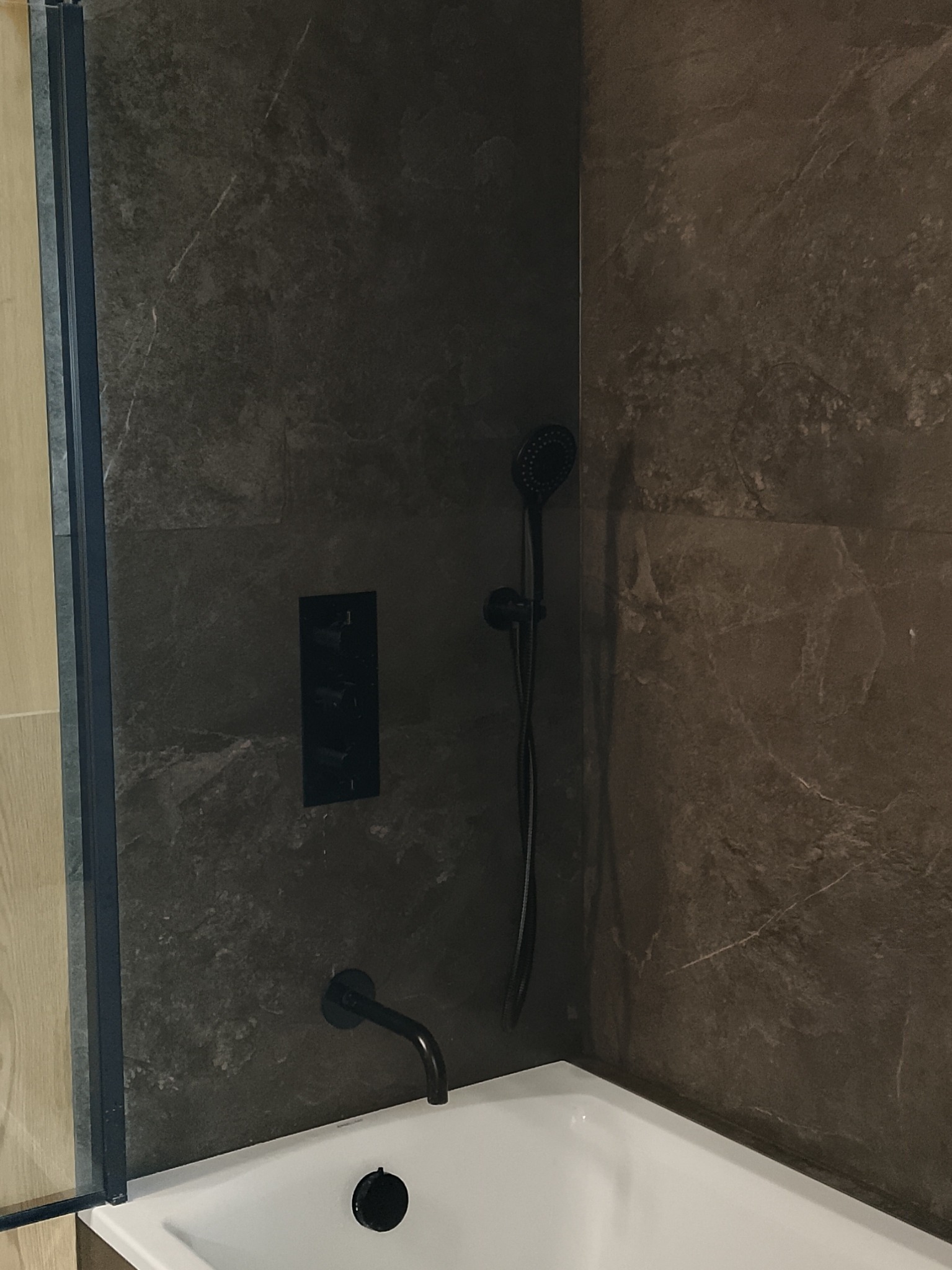 Nowoczesna łazienka z wanną, ciemnymi płytkami imitującymi kamień i czarną armaturą. Widoczny fragment kabiny prysznicowej z drewnianym wykończeniem.