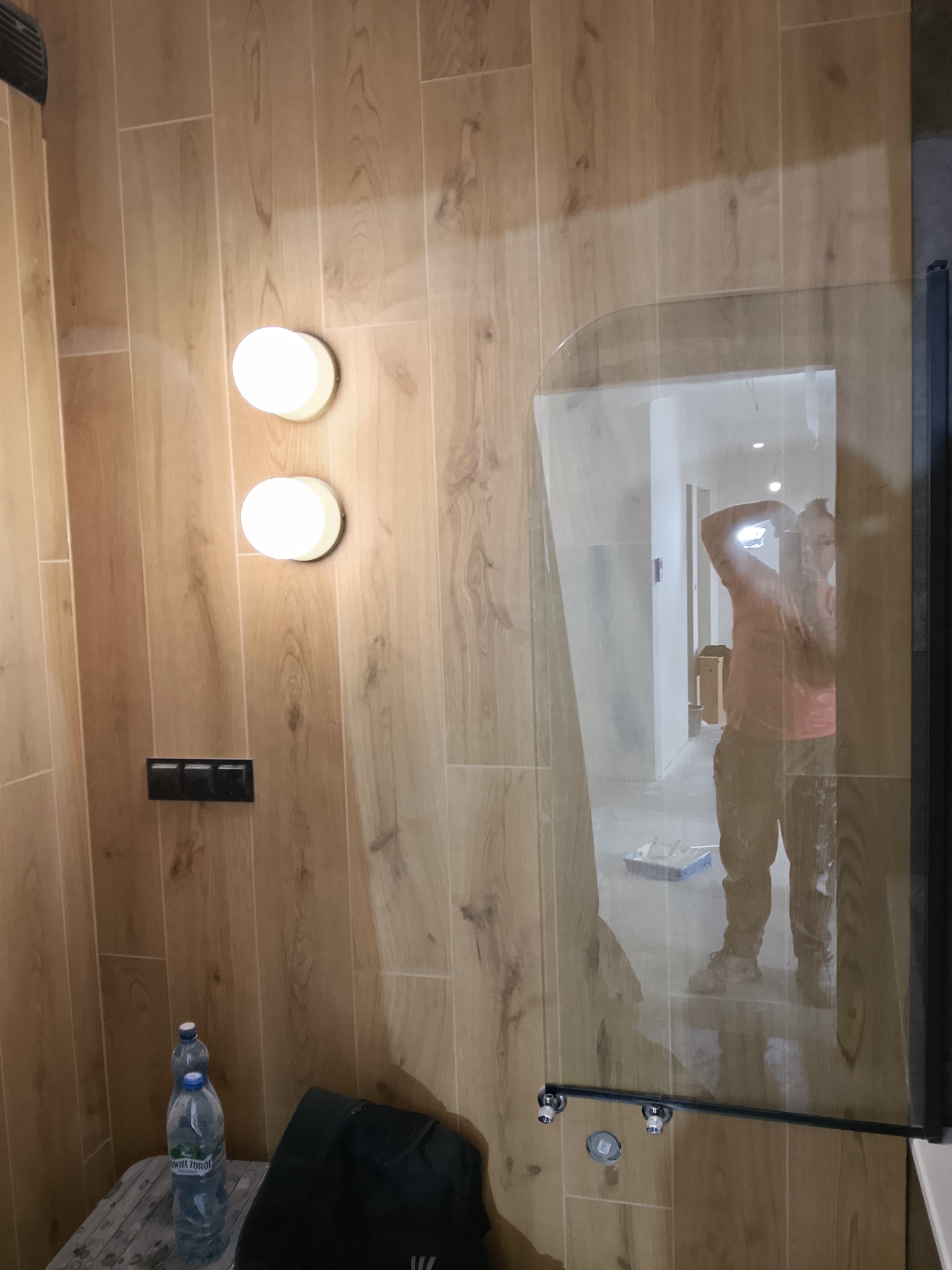 Wykończona łazienka z drewnopodobnymi płytkami, oświetleniem punktowym i szklaną kabiną prysznicową. W lustrze odbija się osoba podczas prac wykończeniowych.