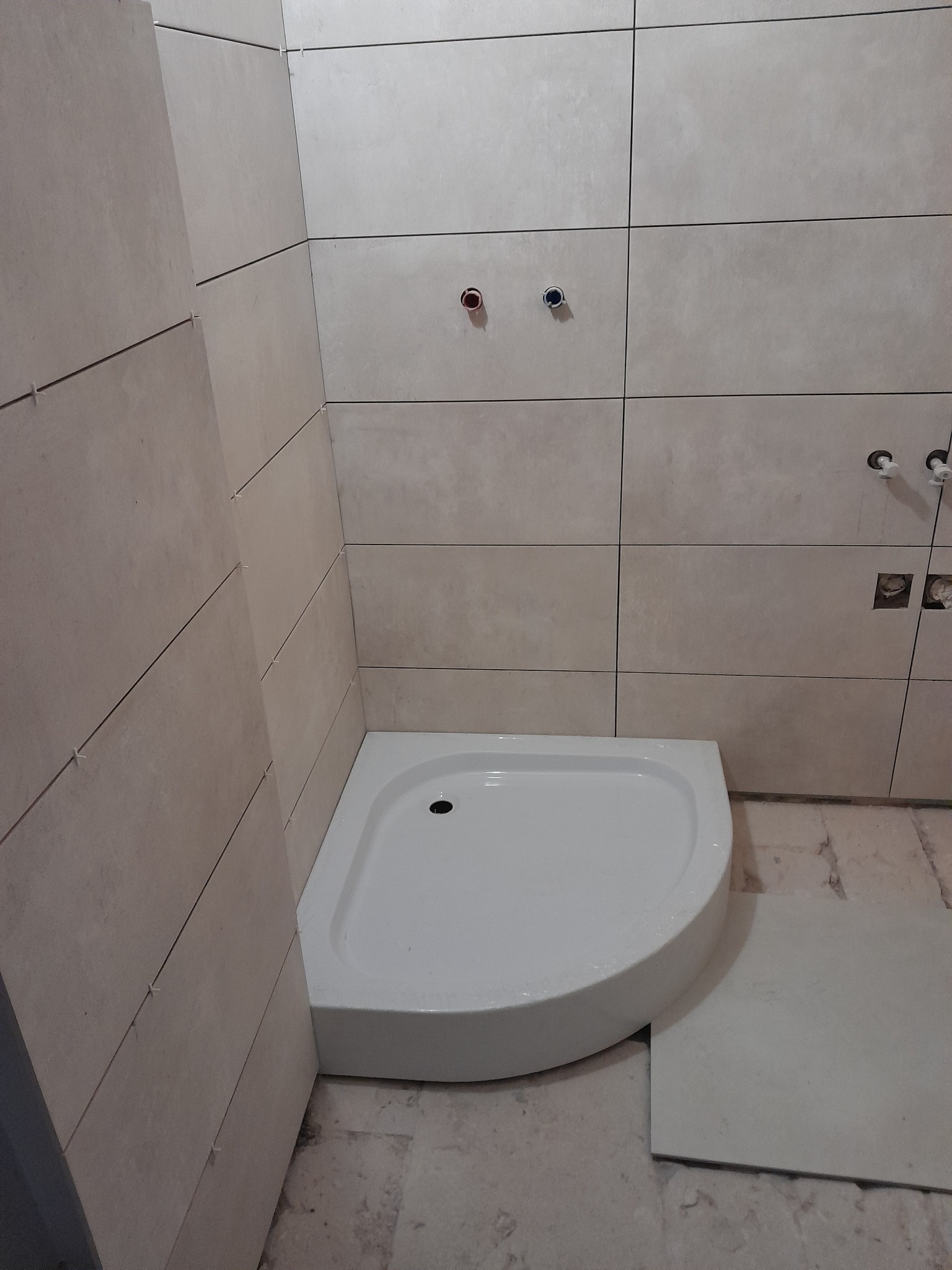 Narożny brodzik w trakcie montażu, obłożony beżowymi płytkami. Widoczne krzyżyki dystansowe i przygotowane punkty instalacyjne na ścianie.