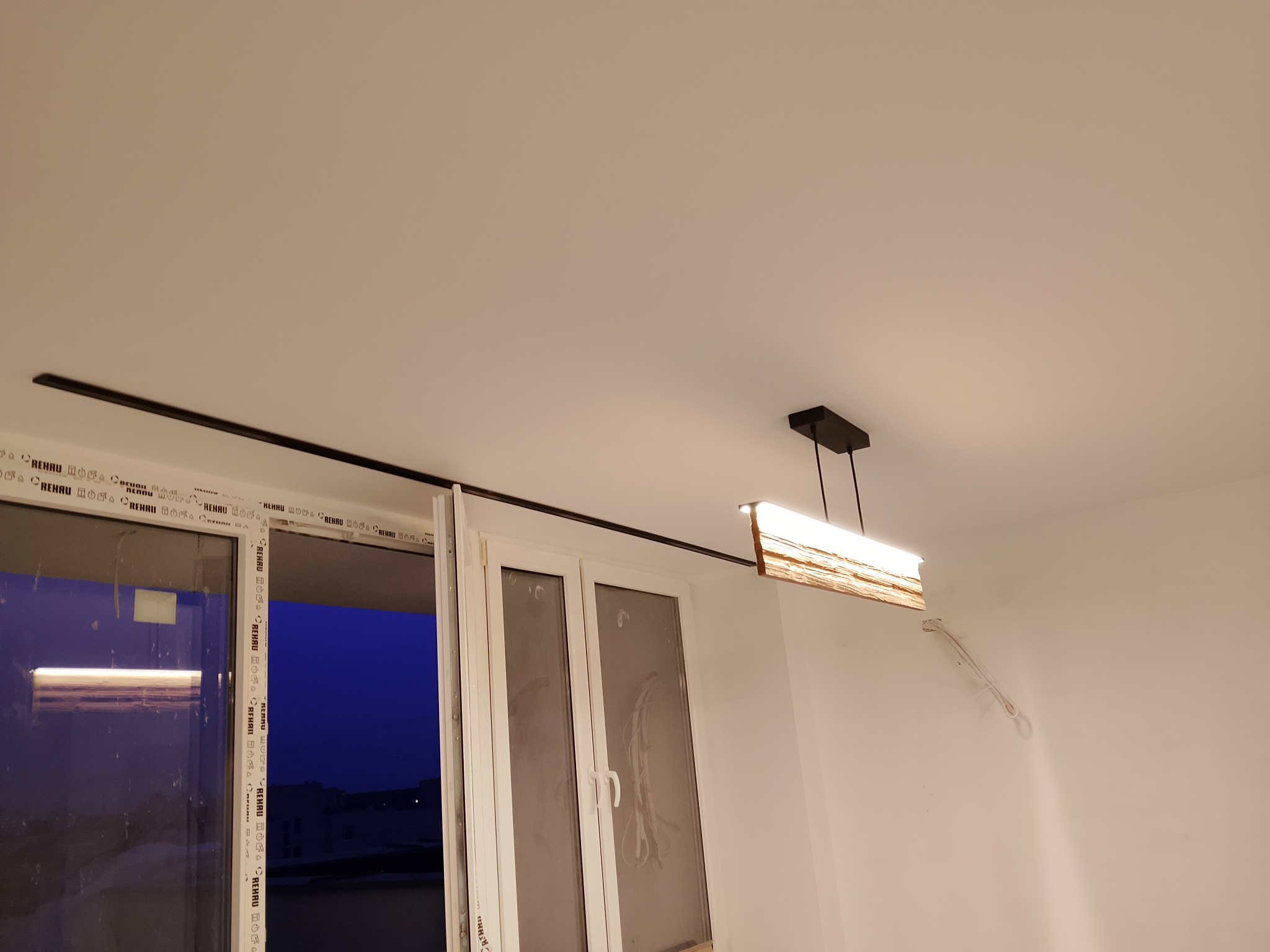 Nowoczesna lampa wisząca z drewnianym akcentem oświetla białe wnętrze z oknem. Widoczne ślady po instalacji elektrycznej na ścianie.