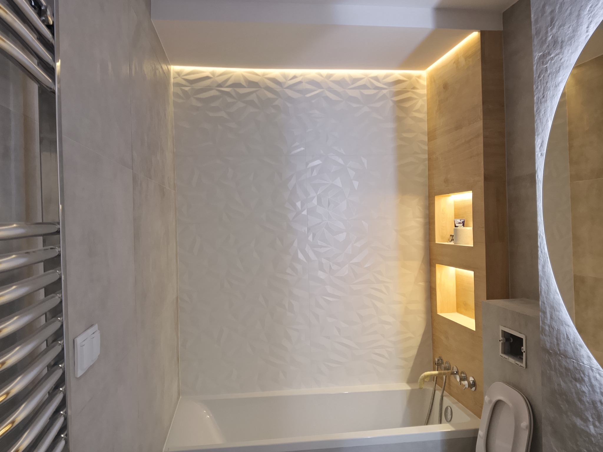 Nowoczesna łazienka z podświetlaną ścianą z trójwymiarowych płytek, wanną i wnękami. Minimalistyczny design z elementami dekoracyjnymi i subtelnym oświetleniem.