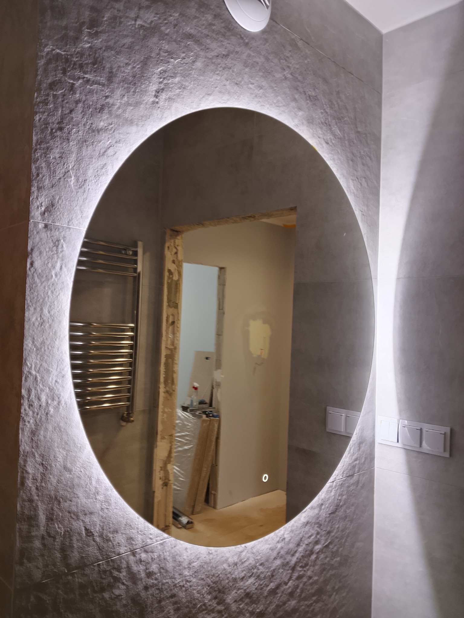 Owalne lustro z podświetleniem LED w nowoczesnej łazience z szarymi płytkami o fakturze kamienia. Widoczny grzejnik drabinkowy i fragment otwartego przejścia do innego pomieszczenia.