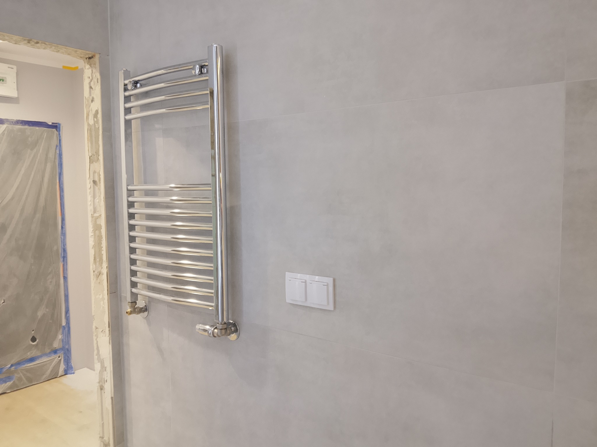 Szary, wykafelkowany fragment łazienki z chromowanym grzejnikiem drabinkowym i podwójnym włącznikiem światła. Widoczny fragment zabezpieczonego folią wejścia.