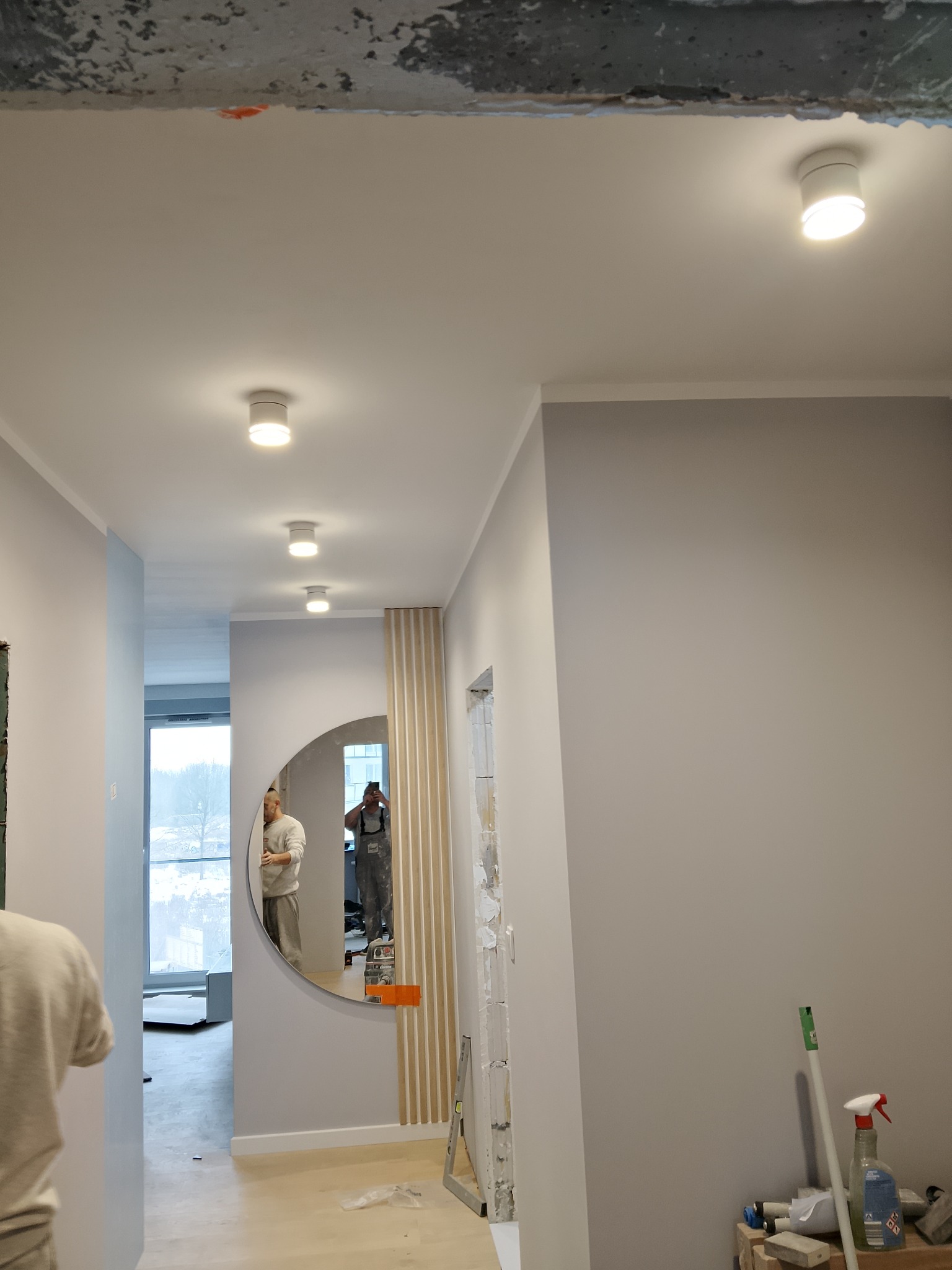 Jasne wnętrze po remoncie: szare ściany, okrągłe lustro z drewnianymi lamelami, oświetlenie punktowe. Widoczne narzędzia i fragment surowego sufitu.