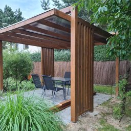 Taras Marzeń - Nowoczesna, brązowa pergola z przezroczystym zadaszeniem, otoczona zielenią ogrodu. Widoczne meble ogrodowe i fragment trawnika.