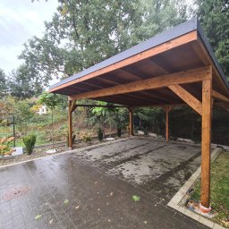 Taras Marzeń - Drewniana wiata garażowa z ciemnym dachem na posesji z kostki brukowej, otoczona zielenią. Widoczne drzewa i krzewy. Konstrukcja z belek w ciepłym odcieniu brązu.