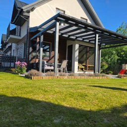 Taras Marzeń - Nowoczesna, czarna pergola z przezroczystym zadaszeniem, przylegająca do domu z balkonem. W tle zielony trawnik i domek dla dzieci. Ujęcie w słoneczny dzień.