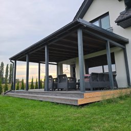 Taras Marzeń - Zadaszony taras z ciemnymi słupami i drewnianą podłogą, otoczony zielonym trawnikiem. W tle widoczny dom z szarym dachem i zielone drzewa.