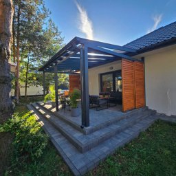 Taras Marzeń - Nowoczesna pergola z drewnianymi panelami bocznymi, przylegająca do domu. Widoczne schody z kostki brukowej i fragment ogrodu. Jasne, słoneczne oświetlenie.