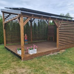 Taras Marzeń - Nowoczesna, otwarta altana z drewnianą podłogą i ażurowymi ścianami, z dzieckiem w środku. Dwa białe kwietniki z różowymi kwiatami na podeście. Tuje w tle.