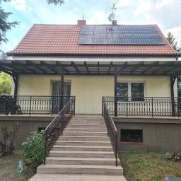 Taras Marzeń - Zadaszony taras z balustradą i schodami wejściowymi, brązowe elementy konstrukcyjne, jasna elewacja domu, panele słoneczne na dachu, otoczenie zieleni.