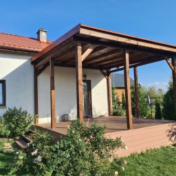 Taras Marzeń - Przydomowa, zadaszona konstrukcja z brązowego drewna, wsparta na słupach, z podestem z desek kompozytowych. Widok na ogród i fragment budynku z oknem.