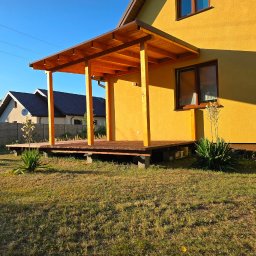 Taras Marzeń - Żółty dom z drewnianym tarasem i zadaszeniem, widok z trawnika. Elementy drewniane w ciepłym odcieniu brązu, kontrastują z kolorem elewacji. Słoneczny dzień.