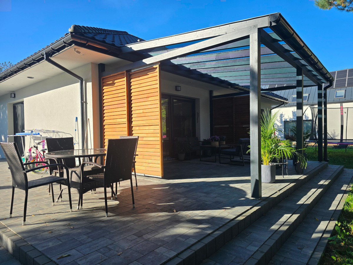 Nowoczesna pergola z ciemnego drewna przylegająca do domu, z drewnianą ścianką działową i meblami ogrodowymi na kamiennym tarasie w słoneczny dzień.