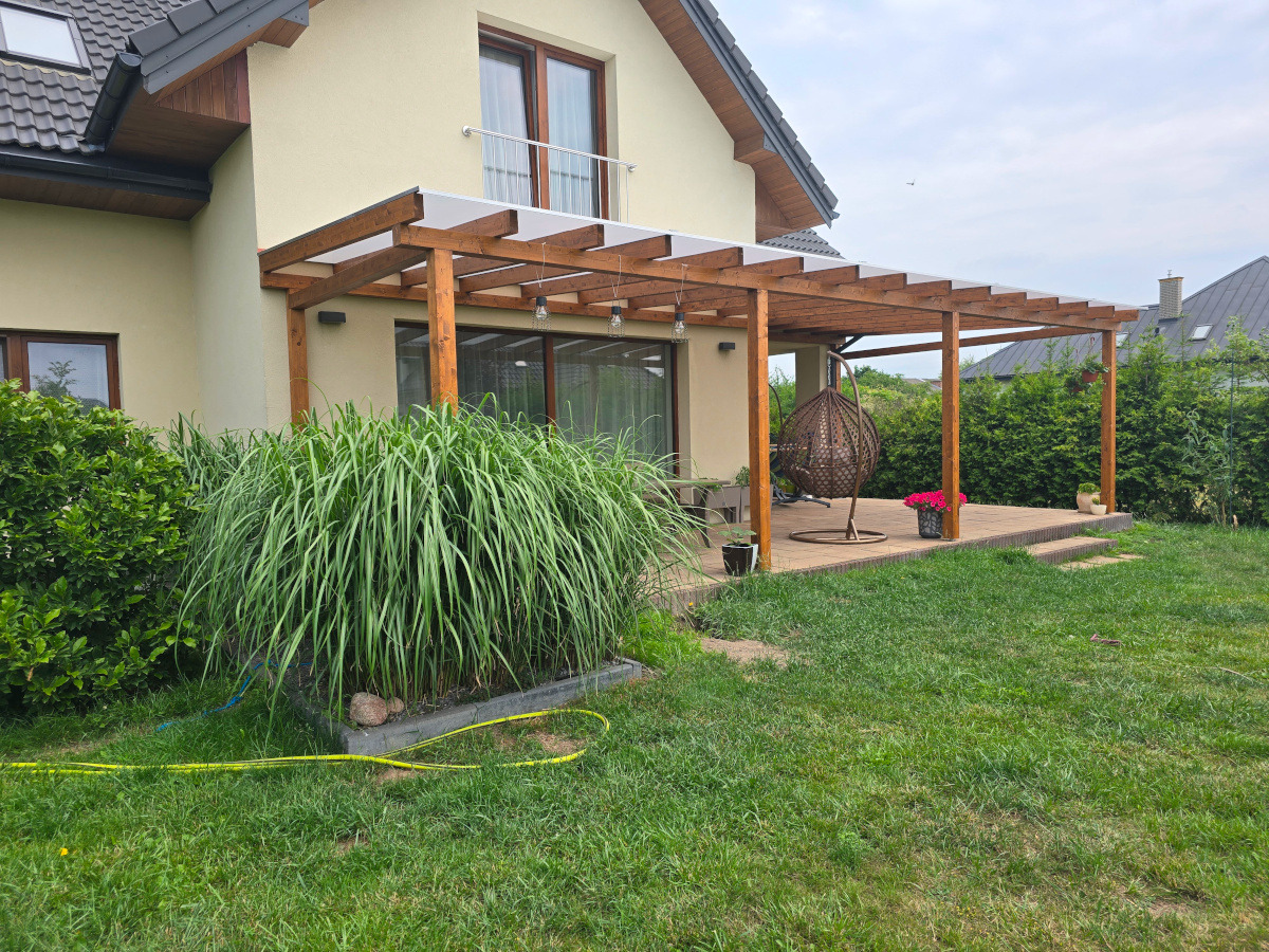 Drewniana pergola z zadaszeniem przy domu, z wiszącym fotelem i roślinami. Widok na taras z kostki brukowej i zadbany trawnik. Oświetlenie dekoracyjne.