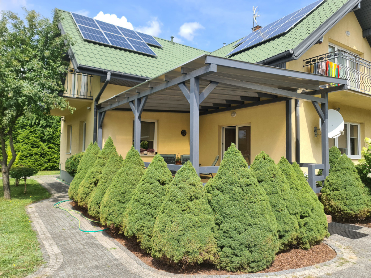 Szara pergola przy żółtym domu z zielonym dachem i panelami słonecznymi, widok z ogrodu z ozdobnymi iglakami i chodnikiem z kostki.