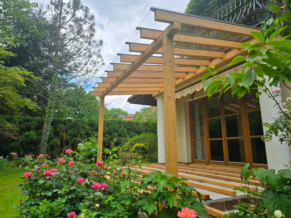 Pergola z przezroczystym zadaszeniem w ogrodzie, widok na konstrukcję z drewna i kwiaty. Etap budowy tarasu z desek. Jasne drewno, zielona roślinność.