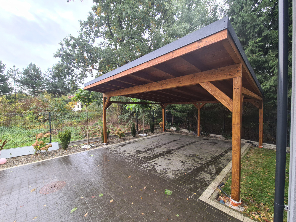 Drewniana wiata garażowa z ciemnym dachem na posesji z kostki brukowej, otoczona zielenią. Widoczne drzewa i krzewy. Konstrukcja z belek w ciepłym odcieniu brązu.