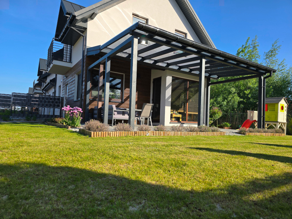 Nowoczesna, czarna pergola z przezroczystym zadaszeniem, przylegająca do domu z balkonem. W tle zielony trawnik i domek dla dzieci. Ujęcie w słoneczny dzień.