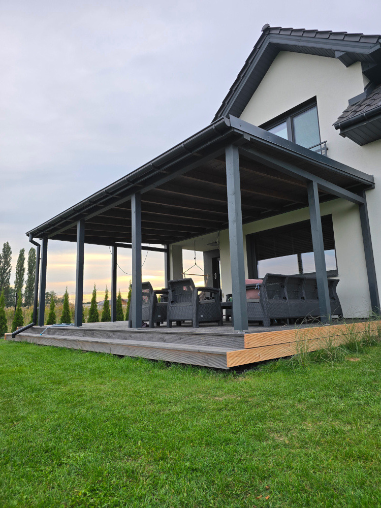 Zadaszony taras z ciemnymi słupami i drewnianą podłogą, otoczony zielonym trawnikiem. W tle widoczny dom z szarym dachem i zielone drzewa.
