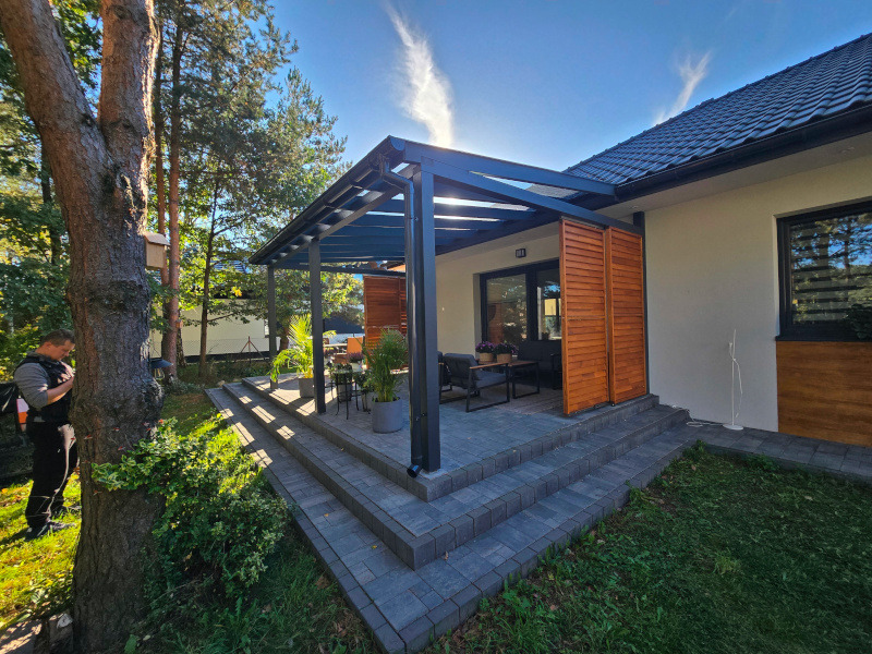 Nowoczesna pergola z drewnianymi panelami bocznymi, przylegająca do domu. Widoczne schody z kostki brukowej i fragment ogrodu. Jasne, słoneczne oświetlenie.