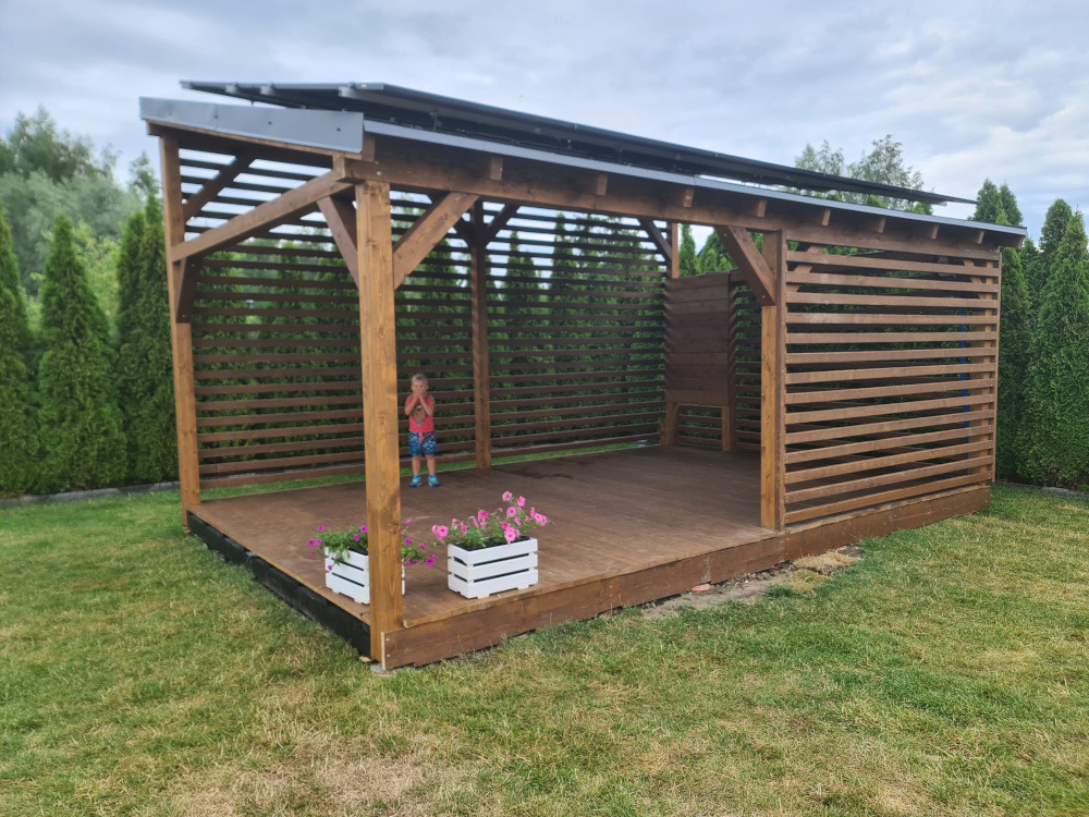 Nowoczesna, otwarta altana z drewnianą podłogą i ażurowymi ścianami, z dzieckiem w środku. Dwa białe kwietniki z różowymi kwiatami na podeście. Tuje w tle.