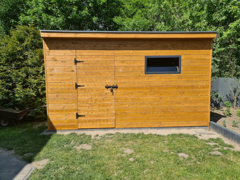 Nowoczesna, drewniana szopa ogrodowa z oknem i czarnymi okuciami, na tle zieleni. Widoczny fragment taczki z lewej strony. Prosta konstrukcja, naturalne wykończenie drewna.