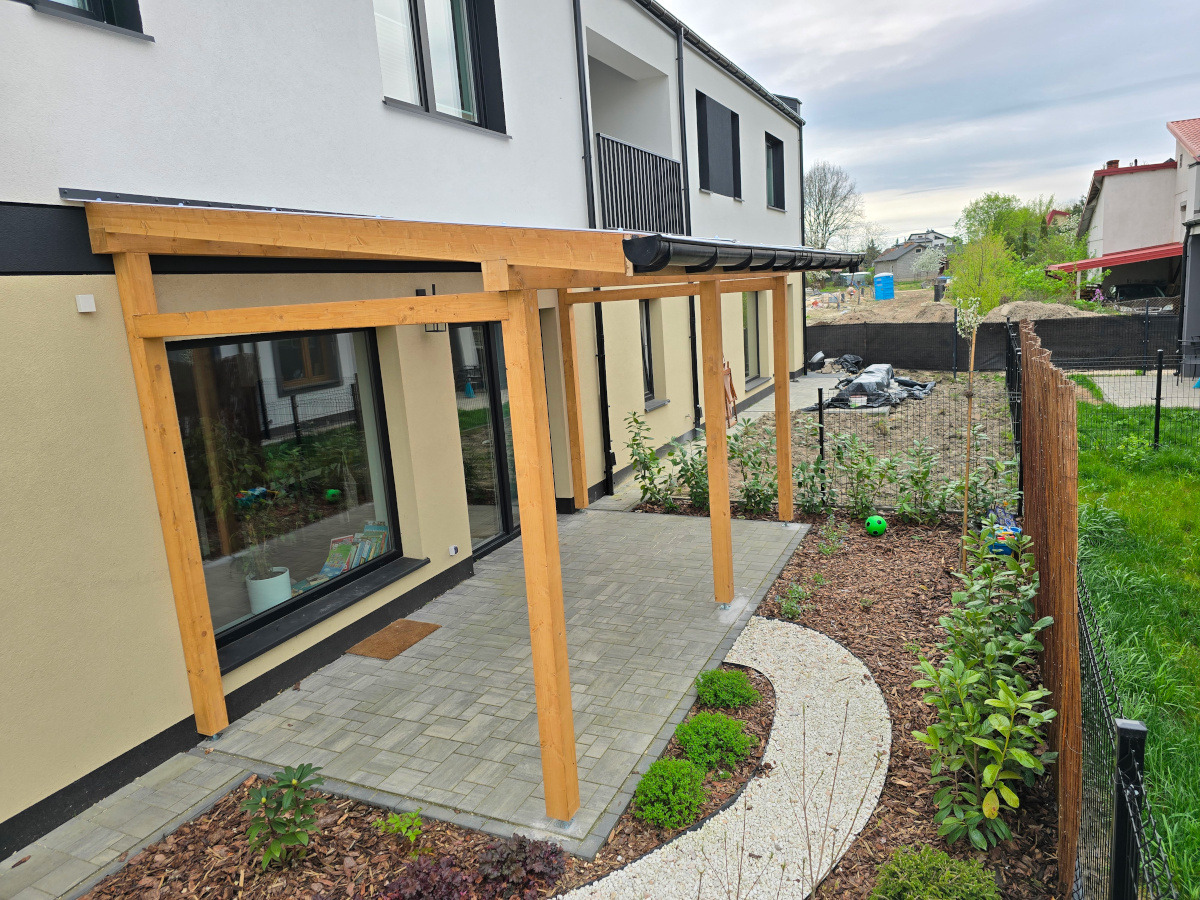 Nowoczesna pergola z jasnego drewna przy domu z szarym brukowanym tarasem i ścieżką z białego żwiru. Widok z góry na zadbany ogród i czarne ogrodzenie.