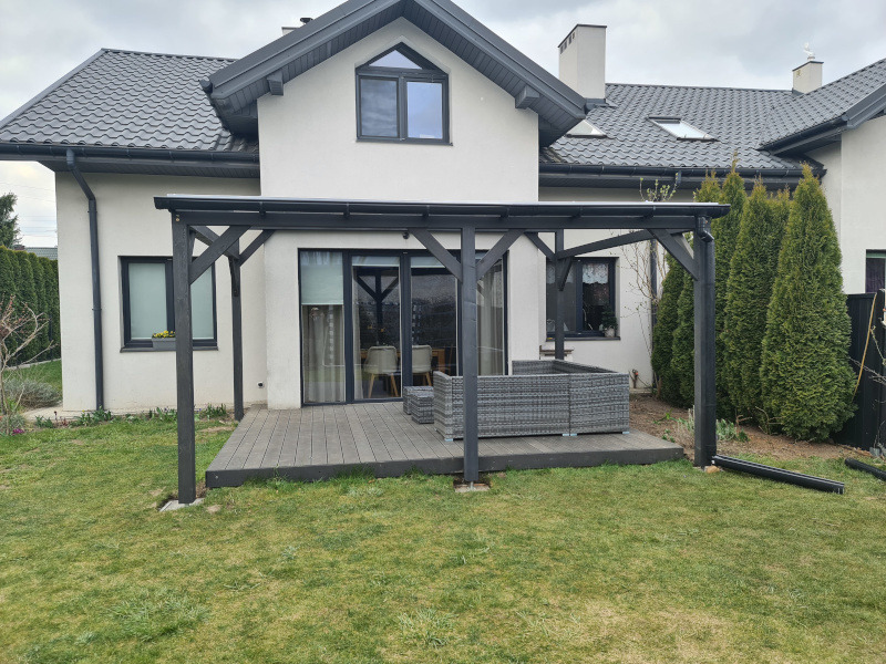 Ciemna pergola drewniana z meblami ogrodowymi na tarasie kompozytowym, przylegająca do białego domu z szarym dachem i zielonym trawnikiem.