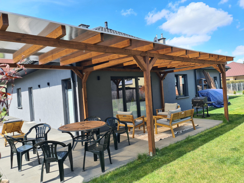 Drewniana pergola z przezroczystym zadaszeniem, z kompletem mebli ogrodowych na tarasie. Widok na szary dom jednorodzinny i zadbany trawnik w słoneczny dzień.