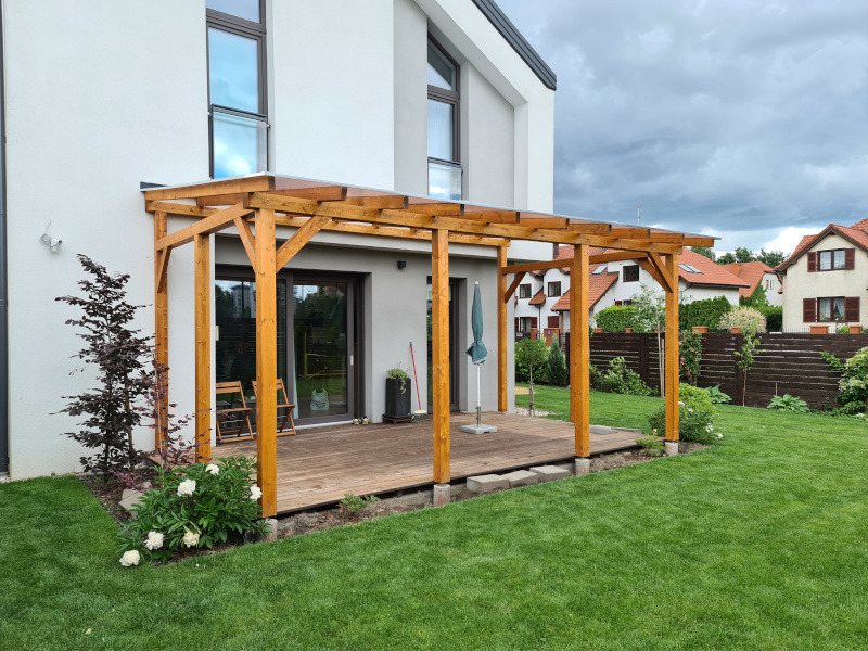 Pergola z drewna na tarasie przy nowoczesnym domu. Widoczne drewniane słupy i zadaszenie, trawnik, krzewy i elementy wyposażenia tarasu.