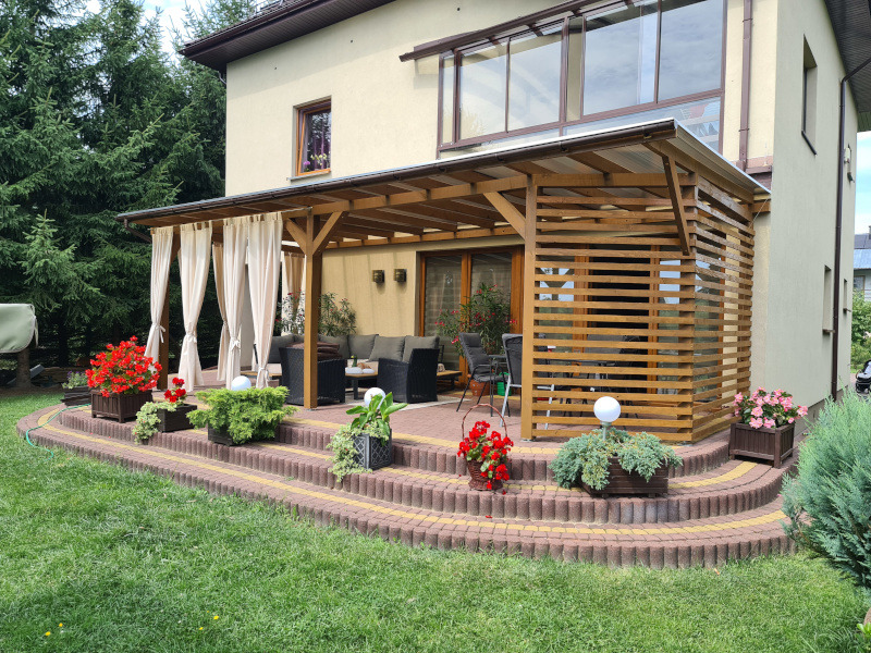 Drewniana pergola z zasłonami i ażurową ścianką, zadaszona, z meblami ogrodowymi na tarasie z kostki, otoczona zielenią. Widok na dom z oknami.