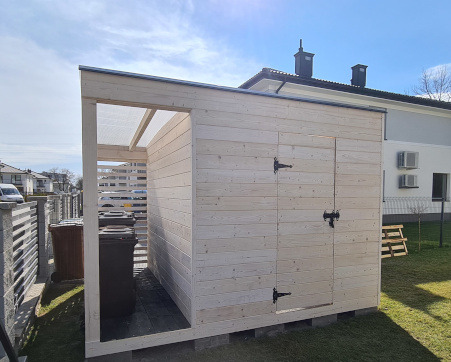 Drewniana konstrukcja z zamykaną częścią i otwartą przestrzenią na pojemniki na śmieci, jasne drewno, widoczne zawiasy, na zewnątrz, w tle dom.