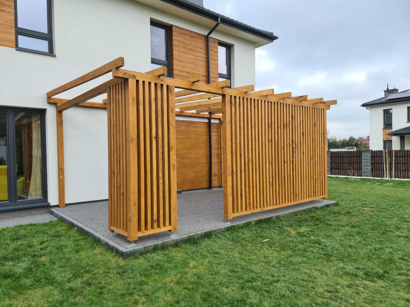 Nowoczesna pergola z drewnianych listew, przylegająca do domu z jasną elewacją. Widoczne staranne wykonanie i dopasowanie do architektury budynku. Szara nawierzchnia.