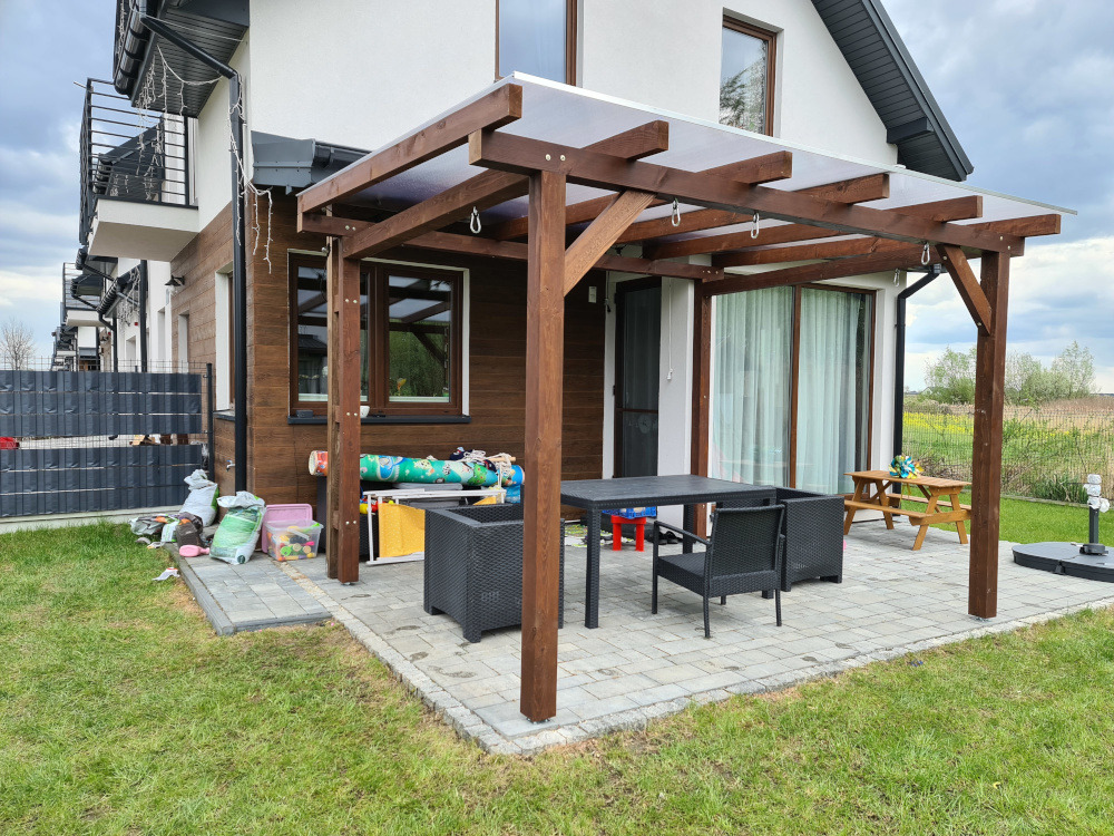 Drewniana konstrukcja z zadaszeniem na kostce brukowej, obok domu z brązową elewacją. Widoczne meble ogrodowe i zabawki. Słoneczny dzień.