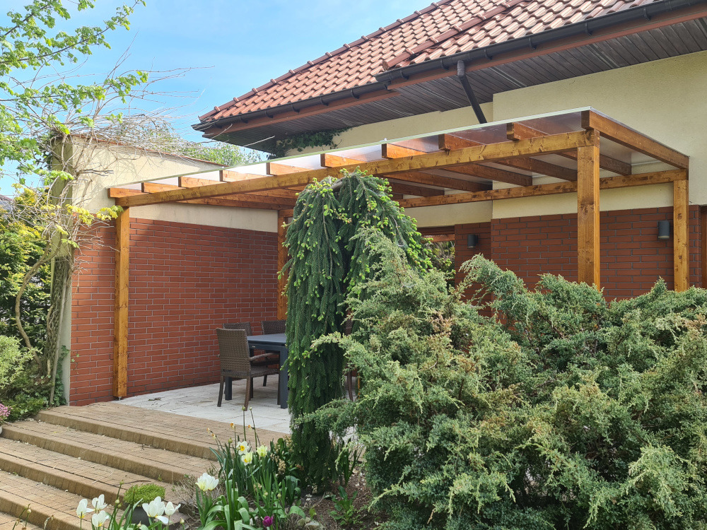 Drewniana pergola z przezroczystym zadaszeniem, przylegająca do ceglanego budynku. W tle stół z krzesłami, a na pierwszym planie bujna zieleń.