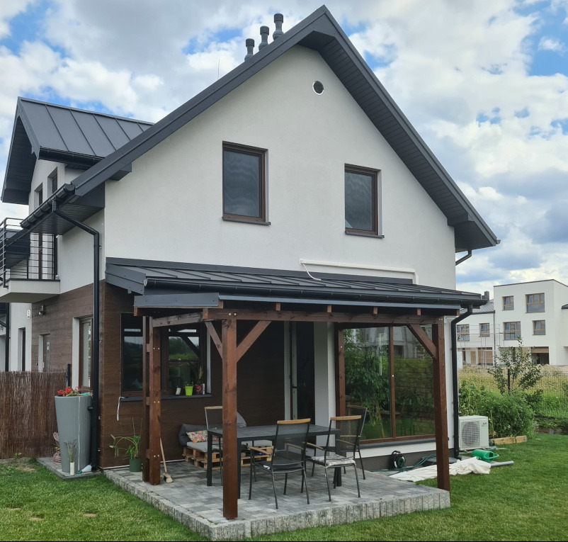 Drewniana konstrukcja zadaszenia tarasu w Komorowie, z widocznym stołem i krzesłami. Ujęcie z perspektywy ogrodu, w tle dom z szarą dachówką i błękitne niebo.