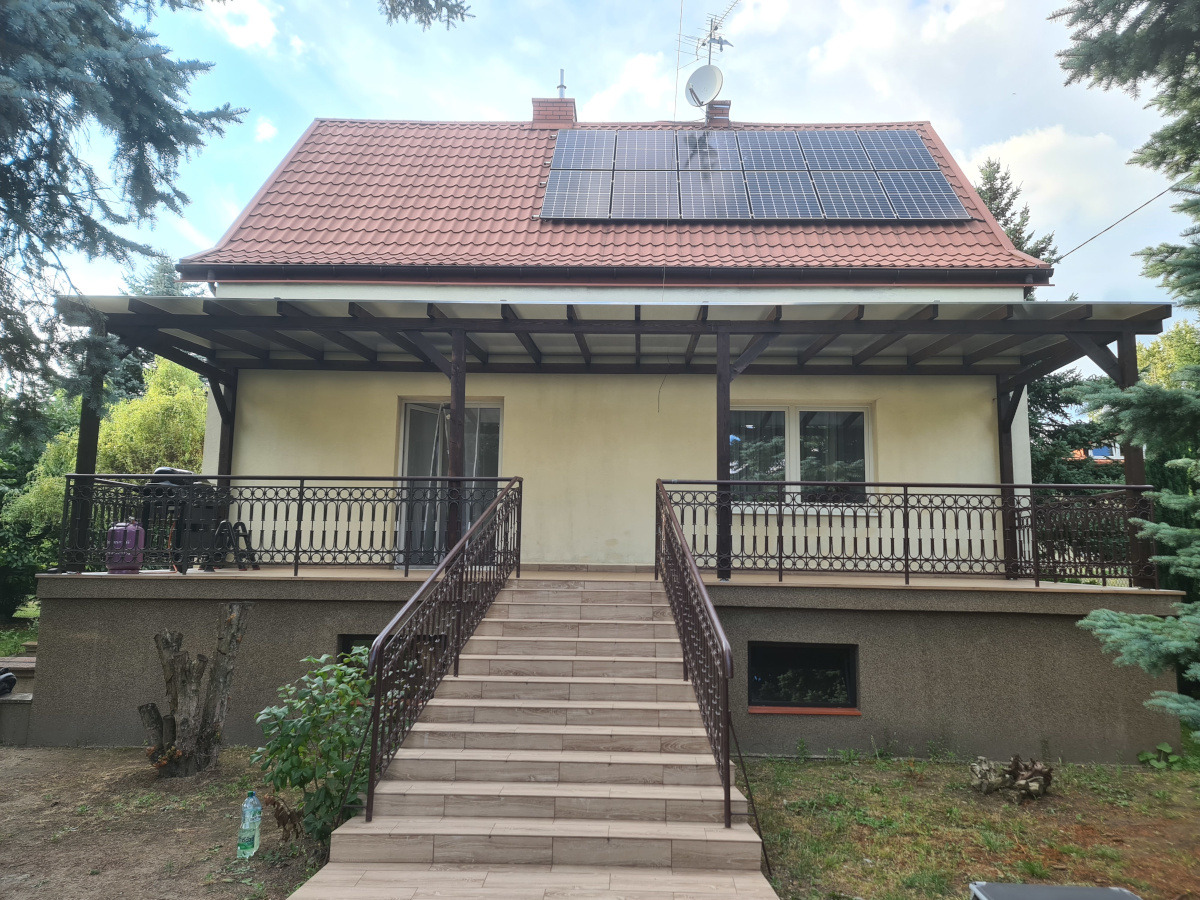 Zadaszony taras z balustradą i schodami wejściowymi, brązowe elementy konstrukcyjne, jasna elewacja domu, panele słoneczne na dachu, otoczenie zieleni.