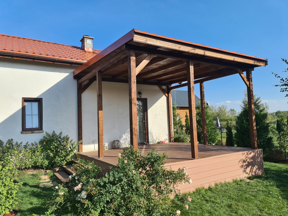 Przydomowa, zadaszona konstrukcja z brązowego drewna, wsparta na słupach, z podestem z desek kompozytowych. Widok na ogród i fragment budynku z oknem.