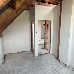 Sar-Bud - Świeżo otynkowane ściany poddasza z widocznymi drewnianymi belkami konstrukcyjnymi dachu i poprowadzonymi przewodami elektrycznymi na betonowej posadzce. W tle otwarte przejścia do kolejnych...