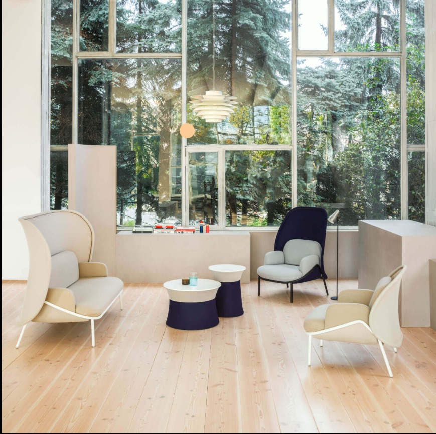 Jasne wnętrze z dużym oknem i widokiem na zieleń. Nowoczesne meble: sofa z wysokim oparciem, krzesła i stoliki w odcieniach beżu i granatu. Drewniana podłoga i designerska lampa.