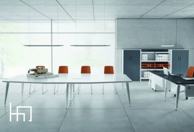 Nowoczesne biuro z długim białym stołem konferencyjnym, pomarańczowymi krzesłami i minimalistycznym oświetleniem. Na stole leży model maszyny.