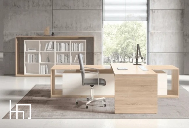 Nowoczesne biuro z biurkiem w kształcie litery L, regałem na książki i krzesłem obrotowym na tle betonowej ściany i okna z widokiem na zieleń. Stonowana kolorystyka.