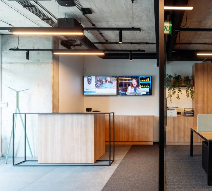 Nowoczesne biuro z recepcją o drewnianym wykończeniu i czarną metalową konstrukcją. Dwa ekrany telewizyjne na ścianie. Widoczne rośliny doniczkowe i betonowy sufit.