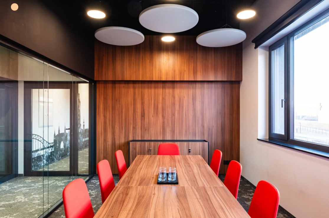 Nowoczesna sala konferencyjna z drewnianą ścianą, czerwonymi krzesłami i okrągłymi lampami sufitowymi. Na stole szklanki z wodą. Za szklaną ścianą widok na miasto.