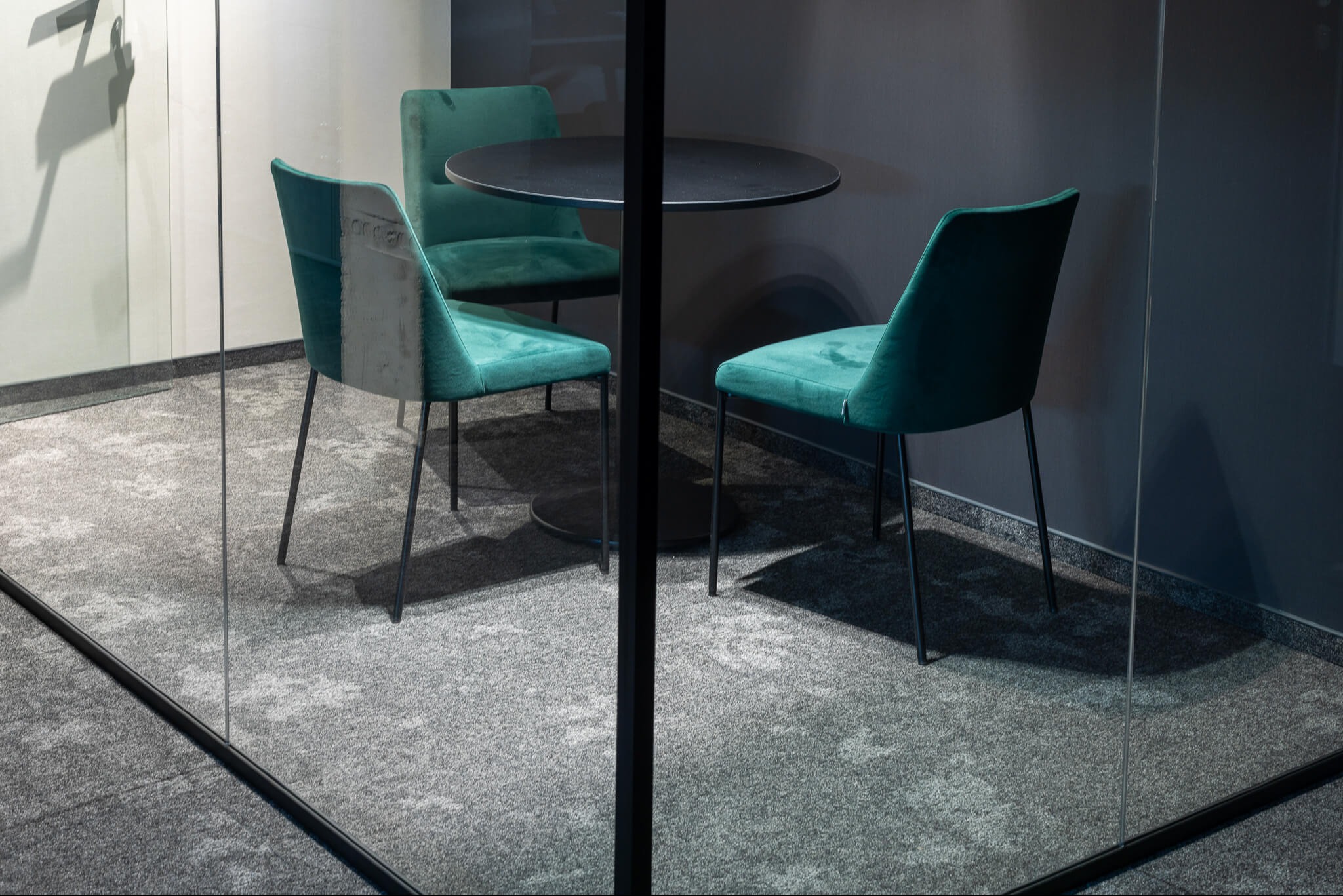 Nowoczesna aranżacja biurowa: okrągły, czarny stół i trzy turkusowe krzesła z weluru w szklanym pokoju. Stonowana kolorystyka, minimalistyczny design.