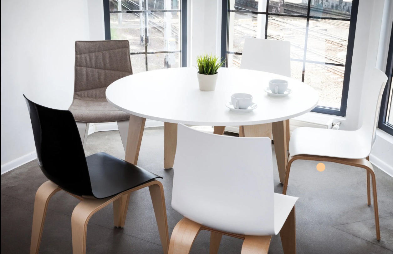 Jasne wnętrze z okrągłym stołem, filiżankami i krzesłami w różnych kolorach. Za oknem widok na tory kolejowe. Minimalistyczny design.