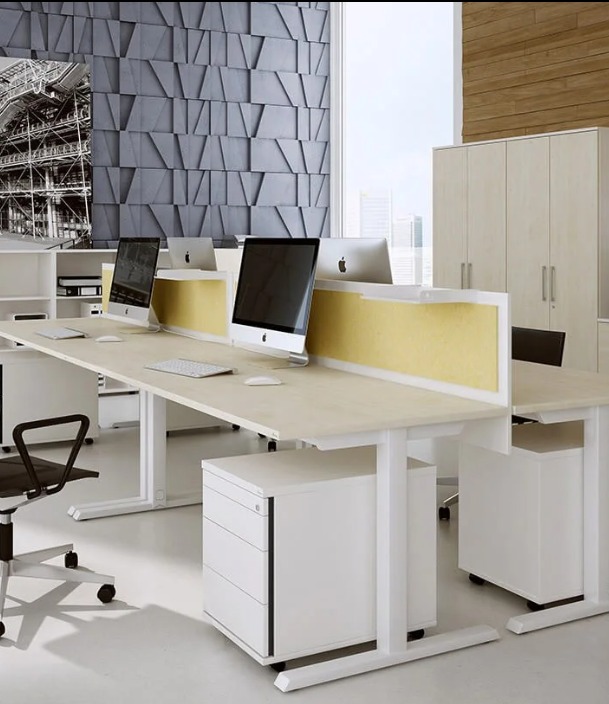 Nowoczesne biuro z długim, jasnym biurkiem, komputerami Apple i szarą ścianą z geometrycznym wzorem. Białe szafki i krzesło biurowe uzupełniają minimalistyczny design.