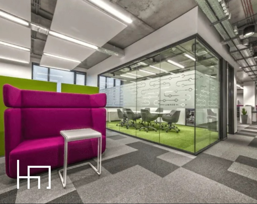 Nowoczesna aranżacja biura: fuksjowa sofa z wysokim oparciem, stolik pomocniczy i przeszklona sala konferencyjna z zieloną podłogą. Geometryczny wzór na wykładzinie.