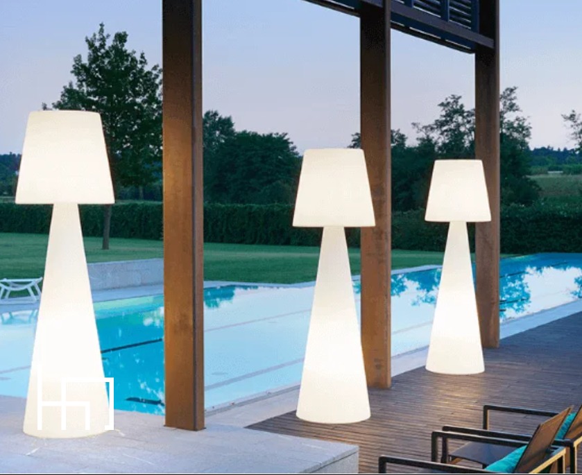 Trzy podświetlane lampy podłogowe w kształcie stożka, oświetlające taras z basenem w tle. Nowoczesny design, ciepłe światło, wieczorny klimat.